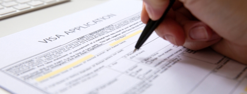 Close up of a female hand filling out a visa application document, white table with computer keyboard and a mouse in background