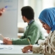 Side view of young serious Muslim woman in blue hijab making notes in copybook while sitting by workplace during presentation